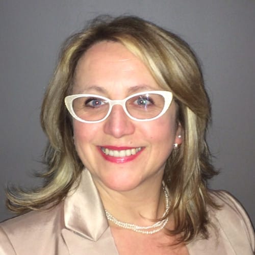 A person with shoulder-length blonde hair, wearing white glasses, pearl necklace, and a beige top, smiles at the camera against a gray background.