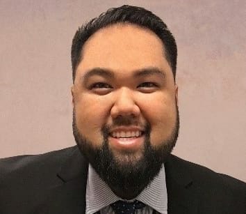 A person with a trimmed beard smiles while wearing a suit and a lanyard, set against a neutral background.