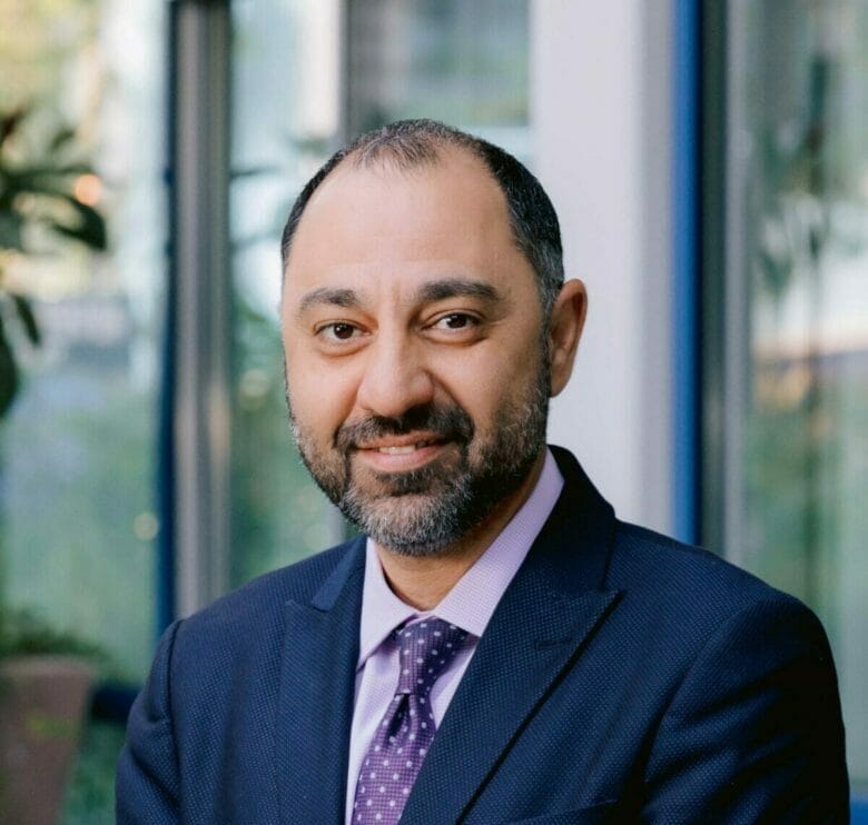 A man with a beard, wearing a dark suit and a purple tie, stands in front of a glass window with plants in the background.