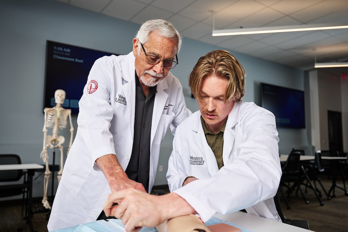 Two people in white lab coats practice a medical procedure on a mannequin arm in a classroom, with a skeleton model in the background.