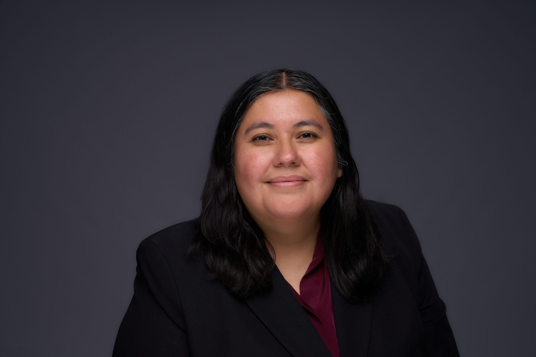A person with long dark hair wearing a dark blazer and maroon shirt sits in front of a plain dark background, facing the camera and smiling slightly.