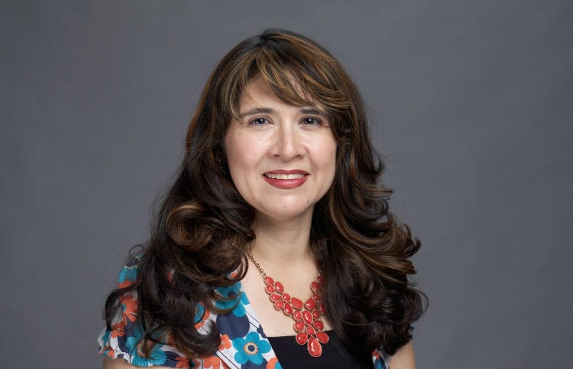 A woman with long brown hair wearing a floral dress, black top, and red necklace poses against a plain gray background.