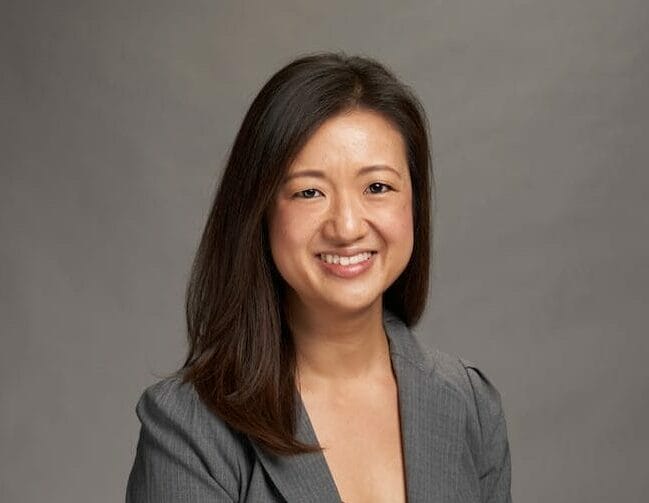 A woman in a gray blazer and dark top smiles at the camera against a plain background.