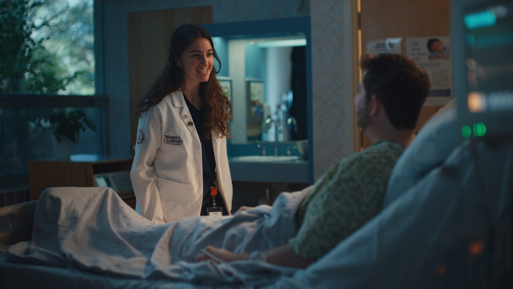 A doctor in a white coat stands by a hospital bed, smiling and talking to a patient who is lying under a blanket in a hospital room.