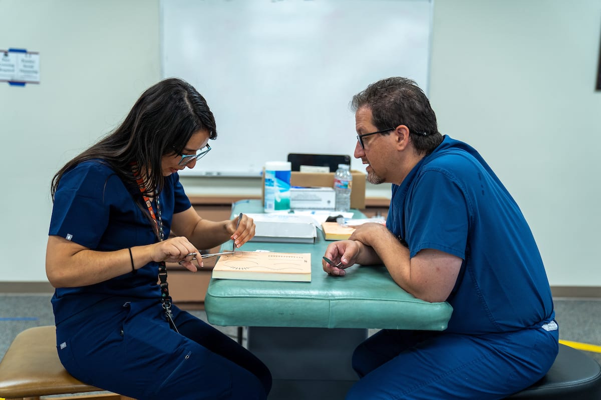 Two people in blue medical scrubs are seated at a table practicing suturing techniques on a simulation pad in a classroom setting.