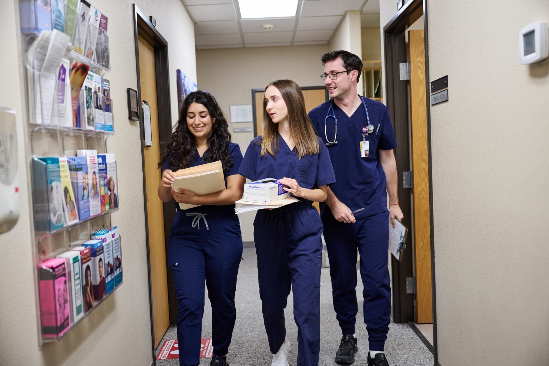 Three healthcare professionals in blue scrubs walk down a hallway, carrying medical supplies and patient files, passing a wall of informational pamphlets.
