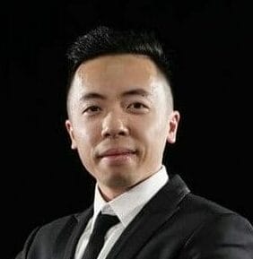 A person in a black suit and tie stands against a dark background with arms crossed.