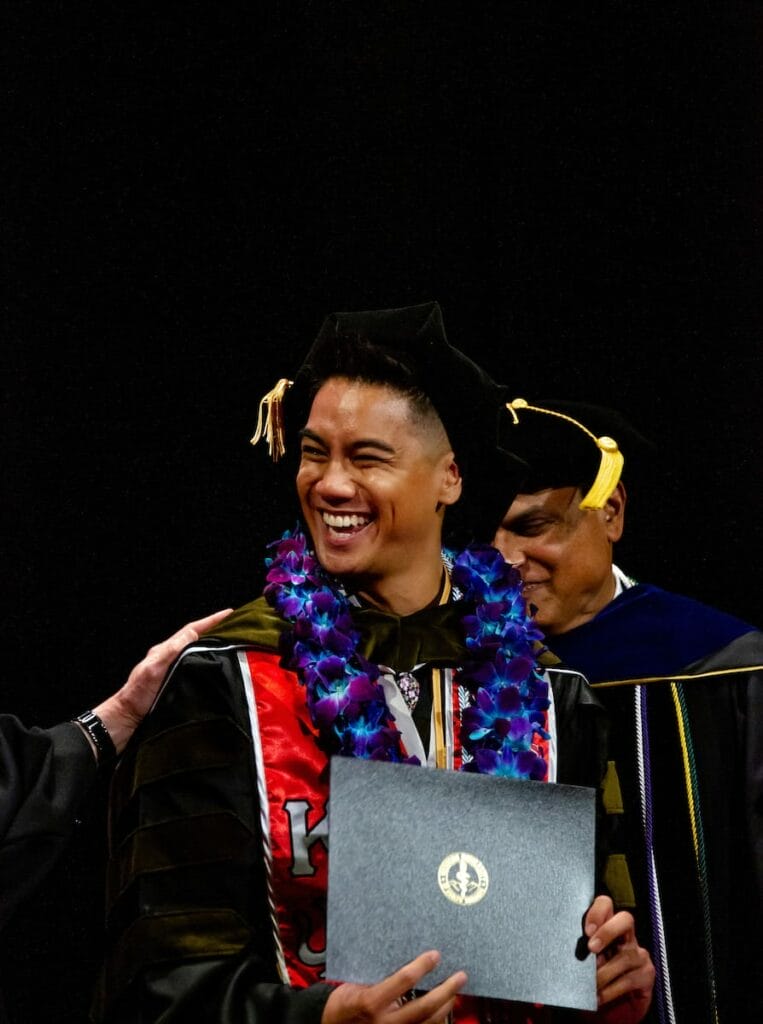 Graduate in cap and gown, adorned with a colorful lei, smiling and holding a diploma on stage, flanked by two faculty members.
