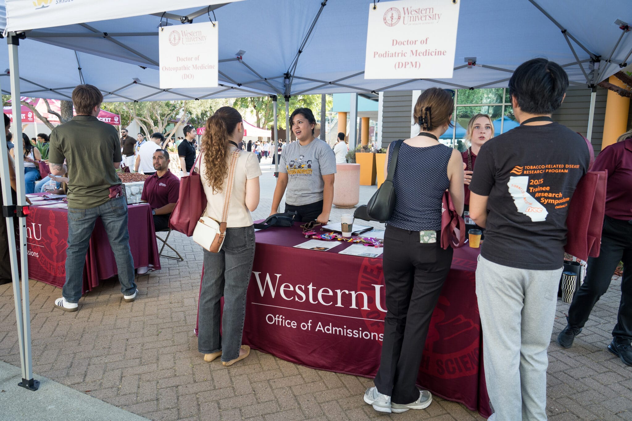 People stand and talk at a Western University Office of Admissions booth for Doctor of Osteopathic Medicine and Doctor of Podiatric Medicine under a tent at an outdoor event.