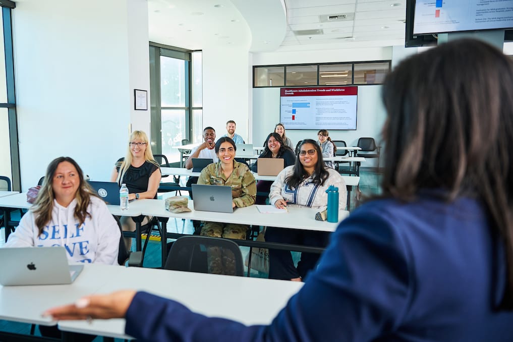 A group of students sit at desks with laptops, facing an instructor at the front of a bright classroom. A presentation slide is projected in the background.