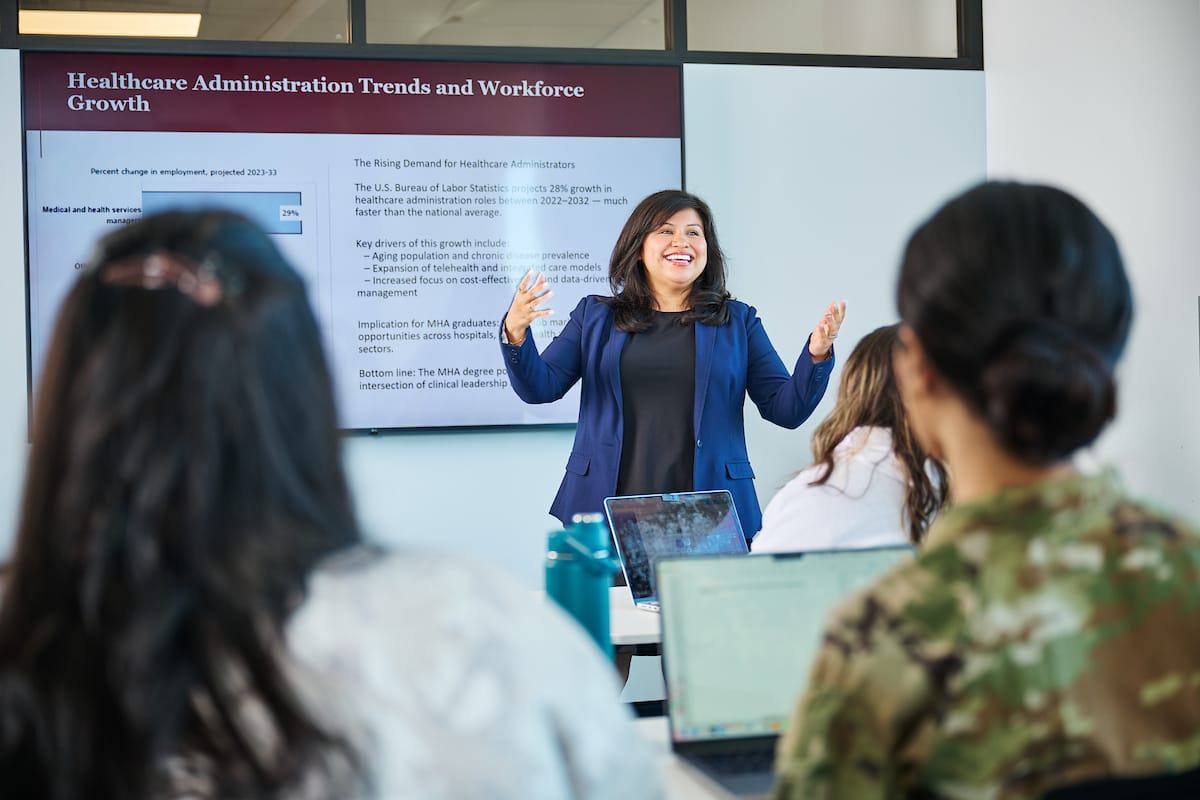 A woman presents healthcare administration trends to a group of students in a classroom; a presentation slide is displayed behind her.