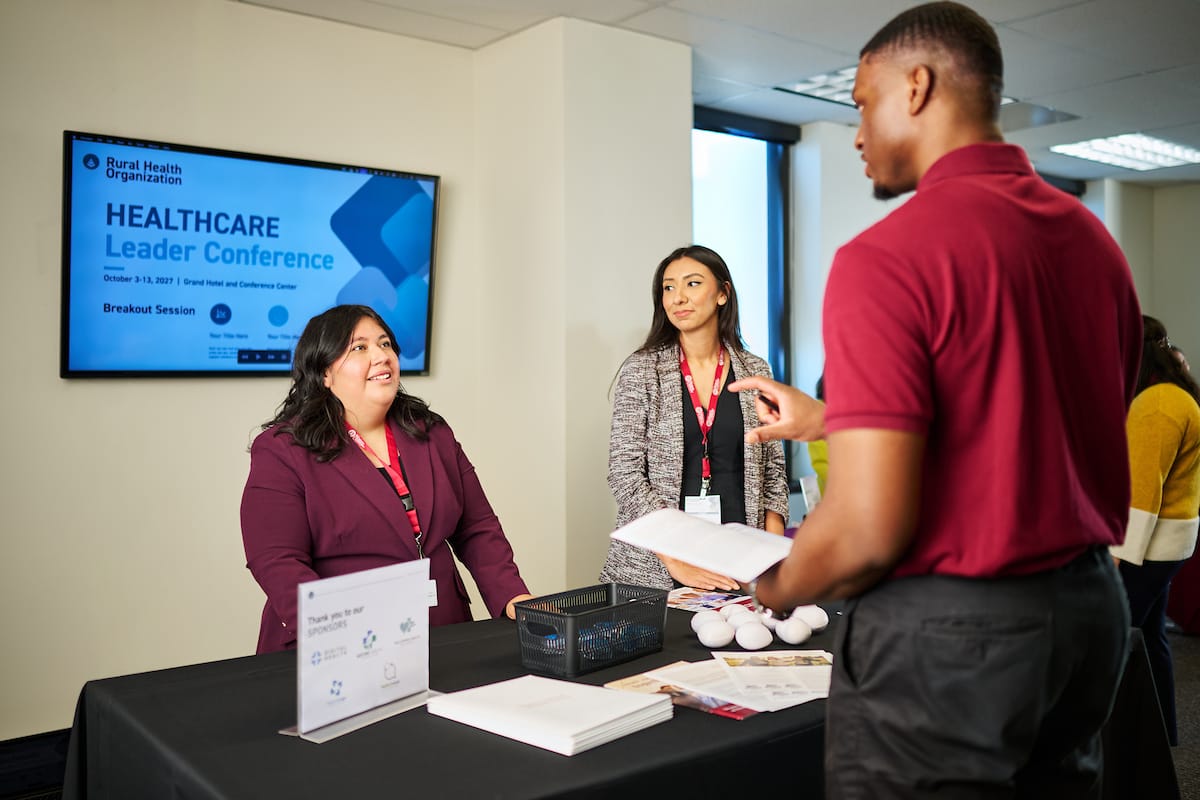Two women stand behind a booth at a healthcare leader conference, talking to a man holding papers; a conference sign is displayed on a screen in the background.