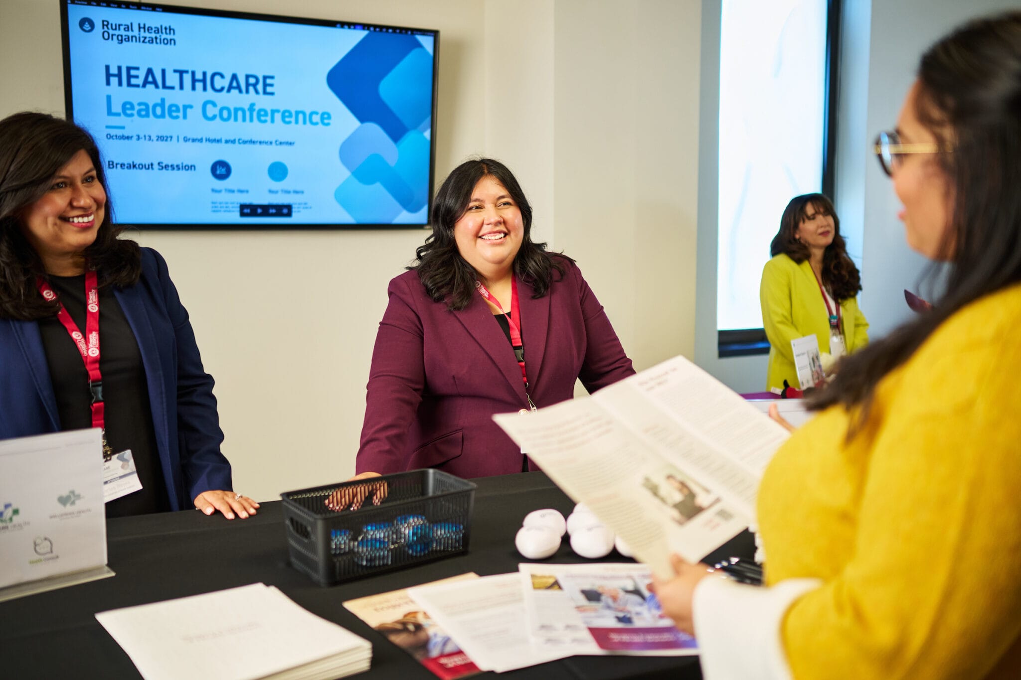 Three women stand at a conference table with brochures, talking to a fourth woman. A screen behind them displays “Healthcare Leader Conference.”.