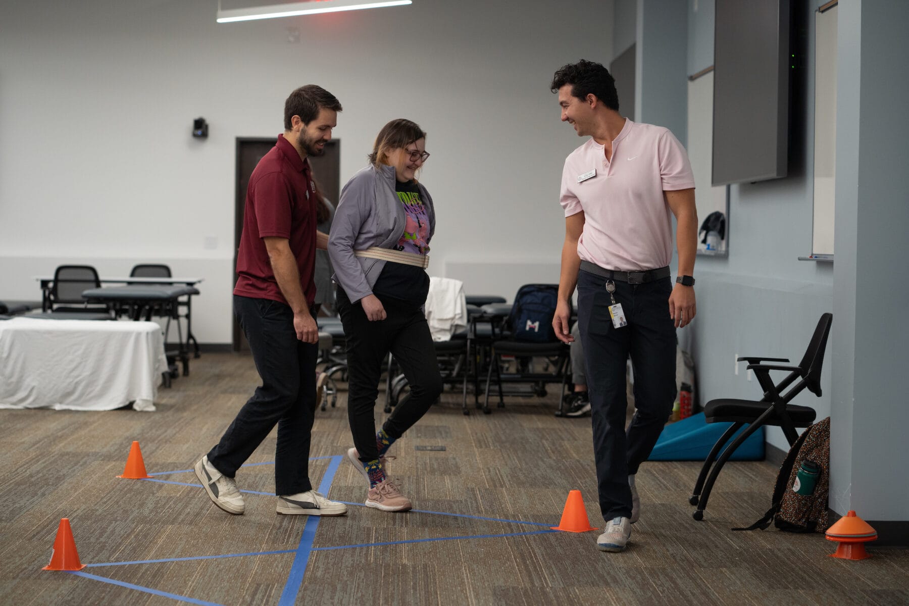 Three people participate in a guided walking activity around orange cones in a classroom setting.