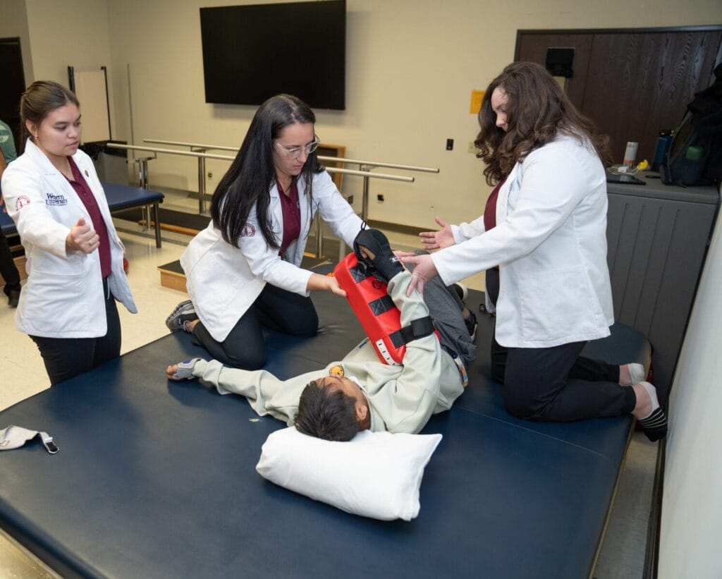 Medical staff in white coats assist a person lying on a treatment table, securing a leg with a brace.
