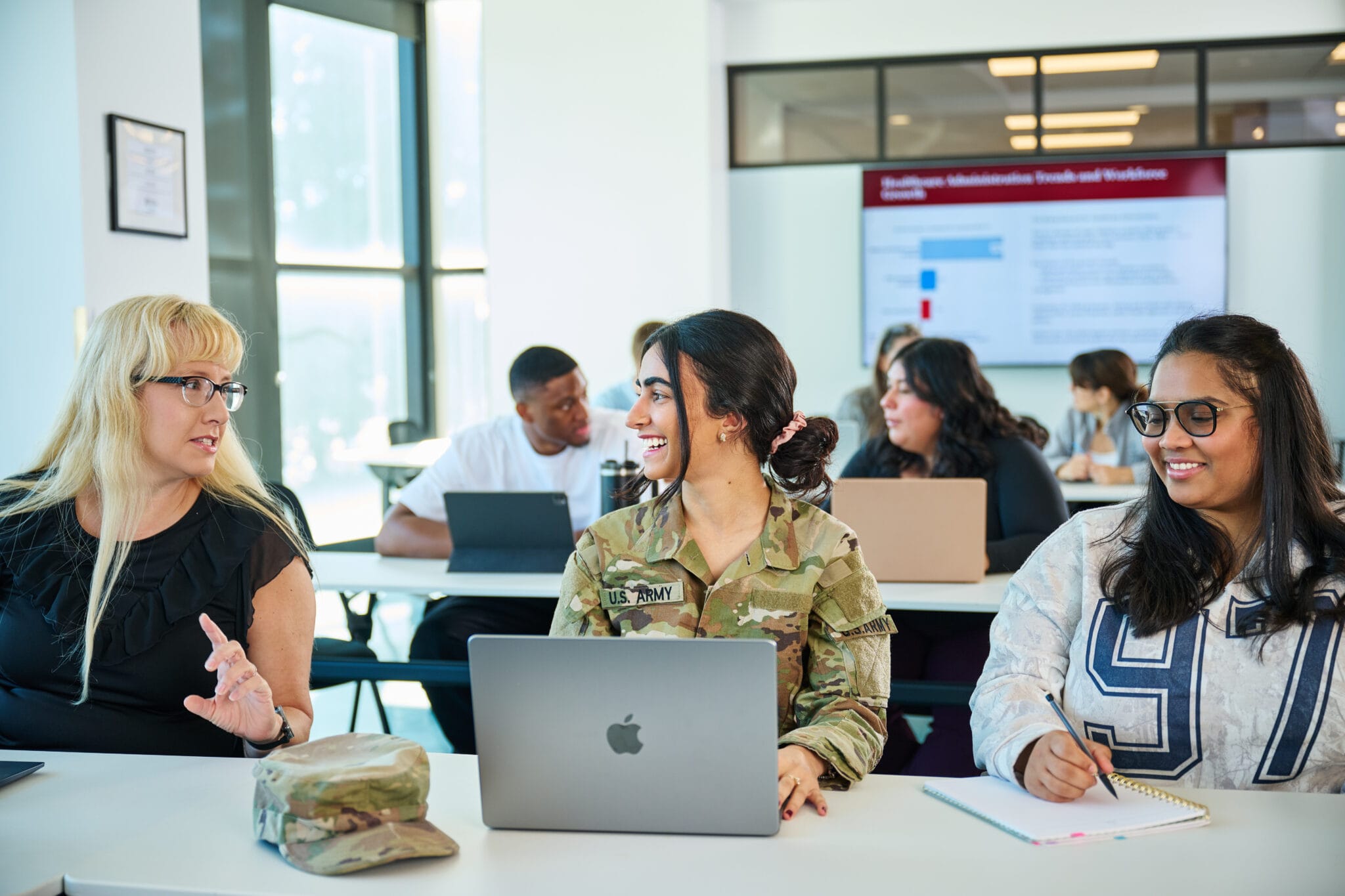 Three women, including one in military uniform, sit at a classroom table with laptops, engaging in conversation; other students work in the background.