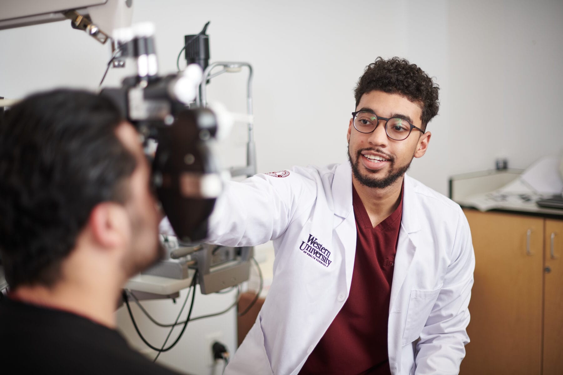 An optometrist in a white coat is examining a patient's eyes using specialized equipment in a clinical setting.