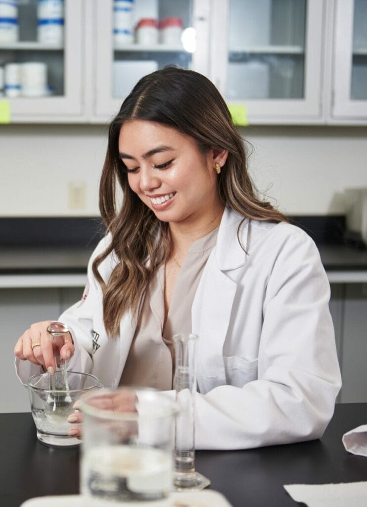 A woman in a lab coat smiles as she conducts an experiment, mixing substances in a bowl with a glass stirrer. Laboratory equipment and glassware are on the table in front of her.