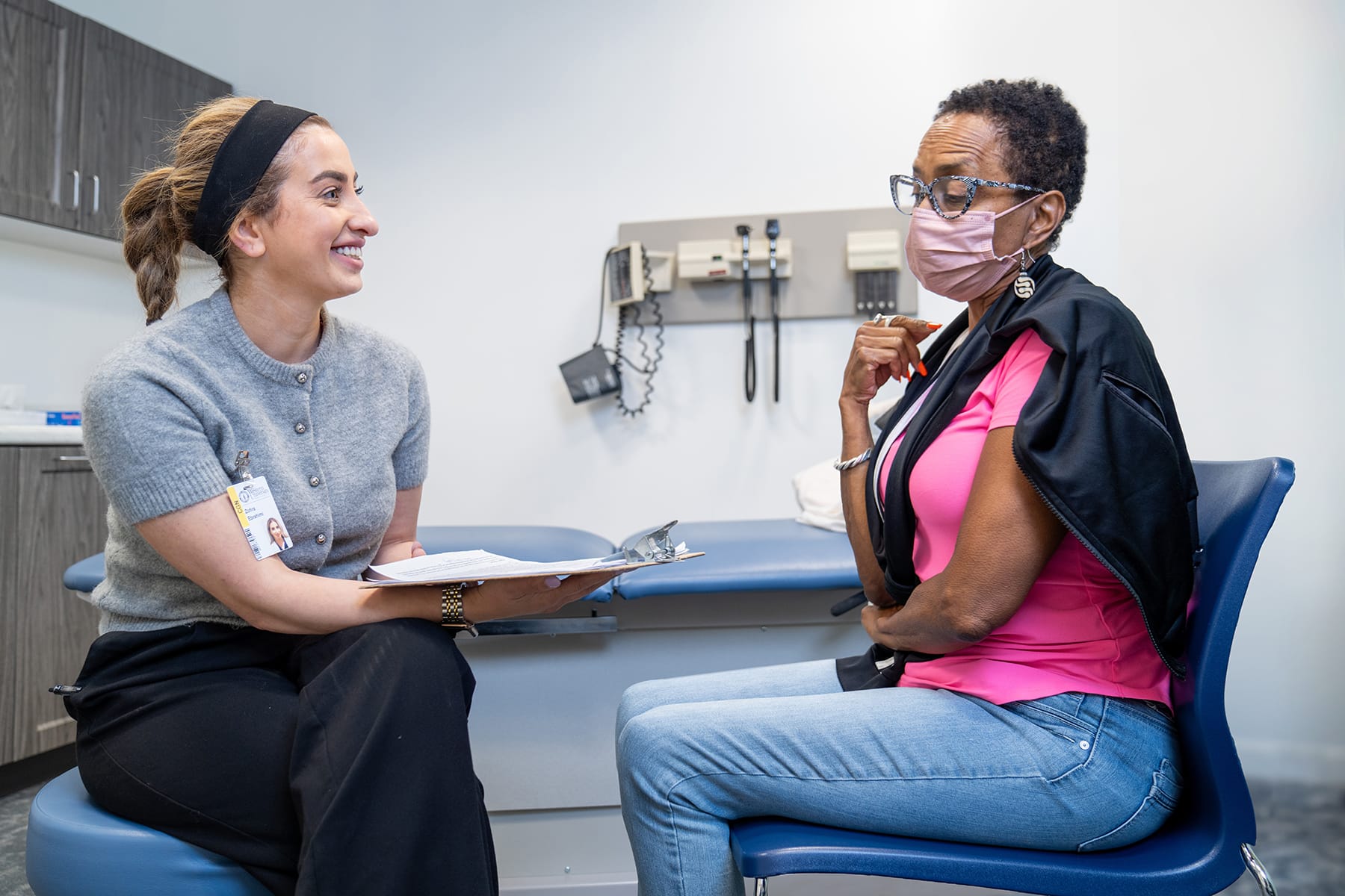 A healthcare professional seated and smiling, holding a clipboard, engages in conversation with a patient wearing a pink shirt and mask, sitting on a chair in a medical examination room.
