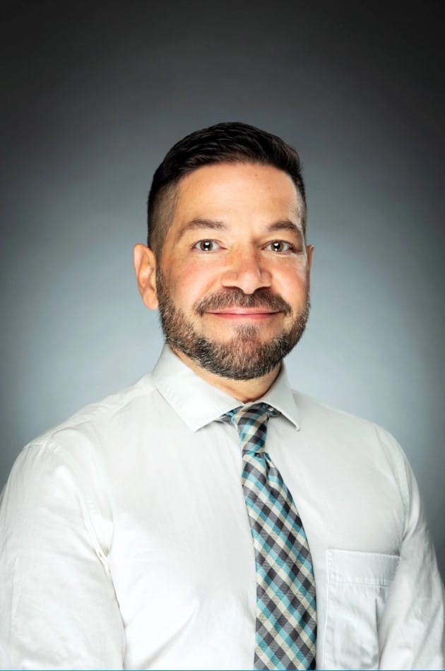 A man with short dark hair and a beard is wearing a white shirt and a patterned tie, posing against a gray background.