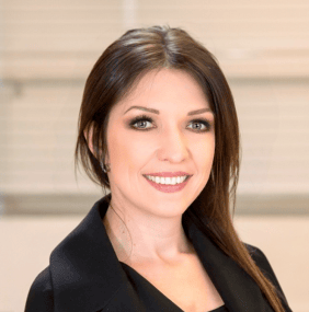 A woman with long brown hair is smiling, wearing a black blazer and standing in front of a blurred background.
