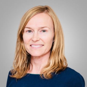 A person with shoulder-length blonde hair and a blue top smiles at the camera against a gray background.