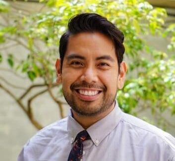 A man with short dark hair and a beard smiles at the camera while wearing a light gray shirt and a floral tie. Green foliage is visible in the background.