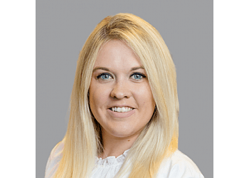Portrait of a woman with long blonde hair smiling, wearing a white shirt, set against a gray background.