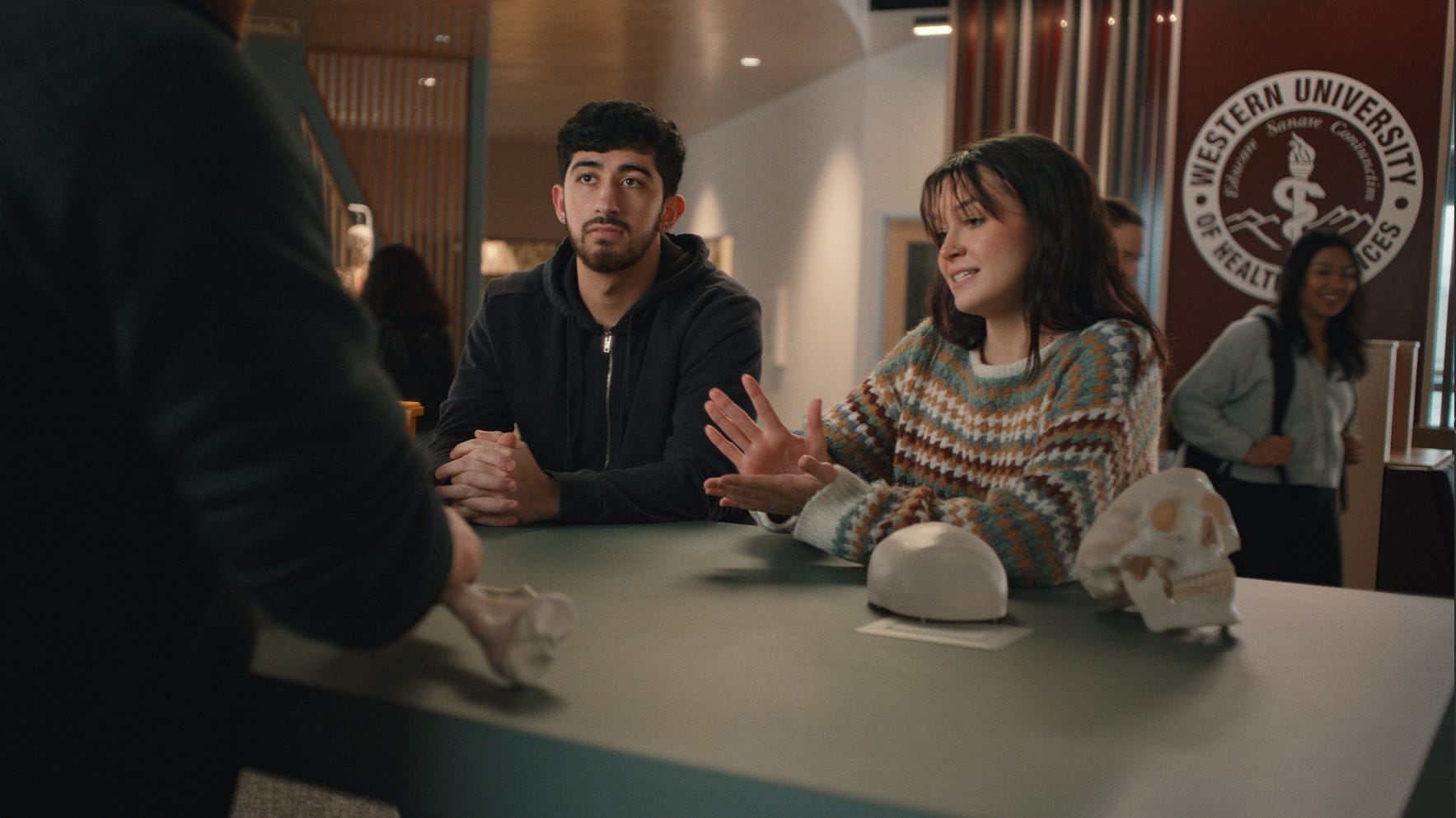 Two students sit at a table with skull models, listening to a person off-camera, inside a building with a "Western University of Health Sciences" sign in the background.