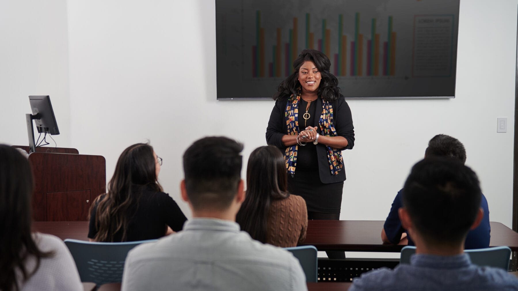 A woman stands in front of a group of seated individuals, giving a presentation. A screen behind her displays a bar chart.