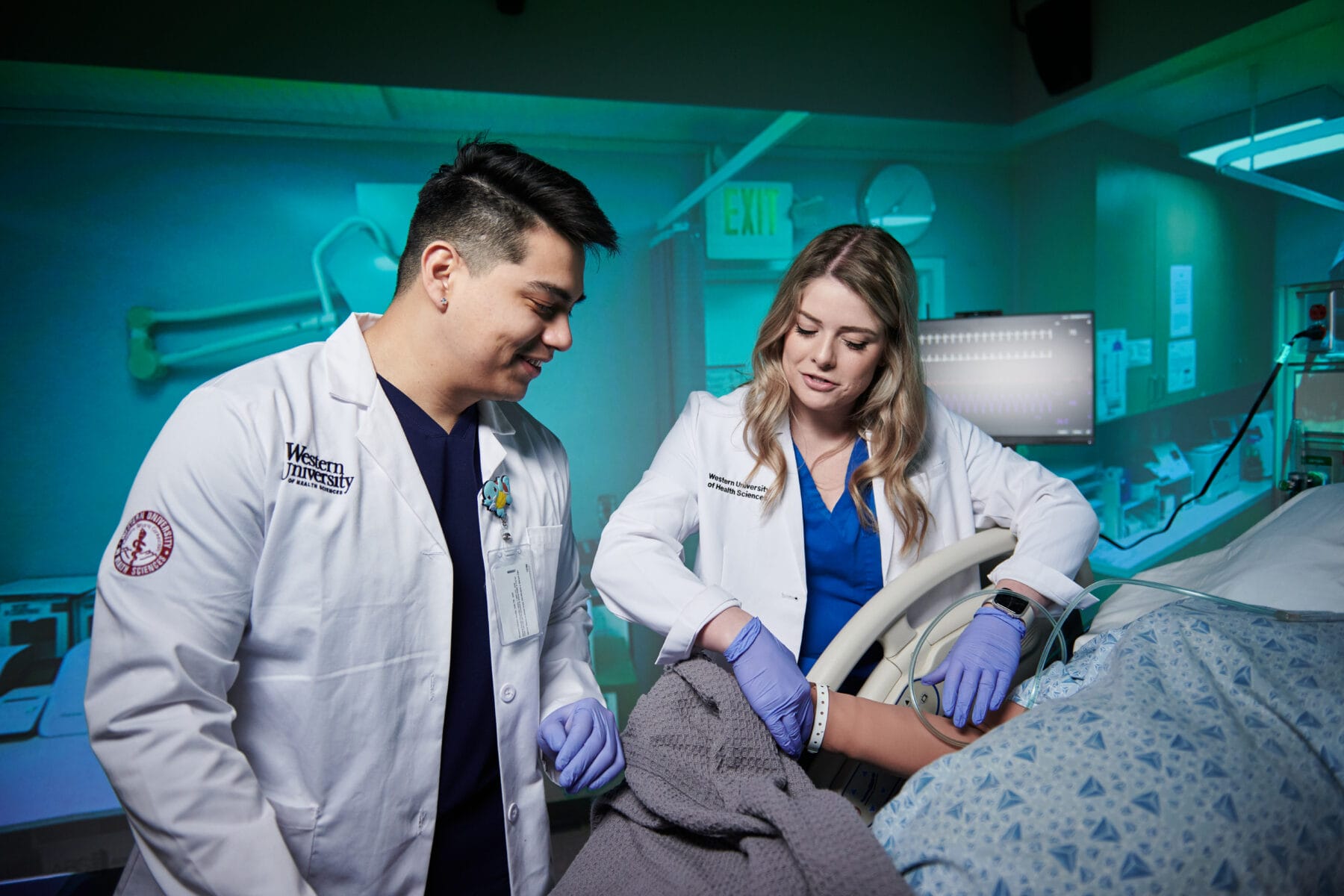 Two medical professionals in lab coats and gloves examine a patient in a hospital room with medical equipment.