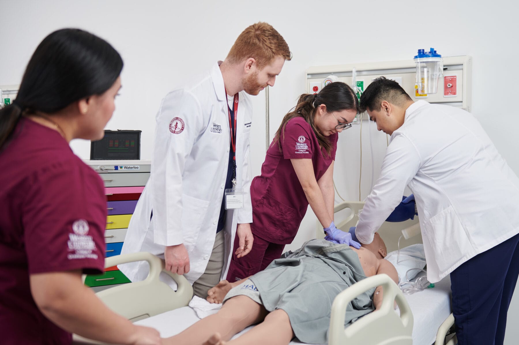 Four medical professionals, including a person performing chest compressions, are surrounding and treating a patient in a hospital bed.