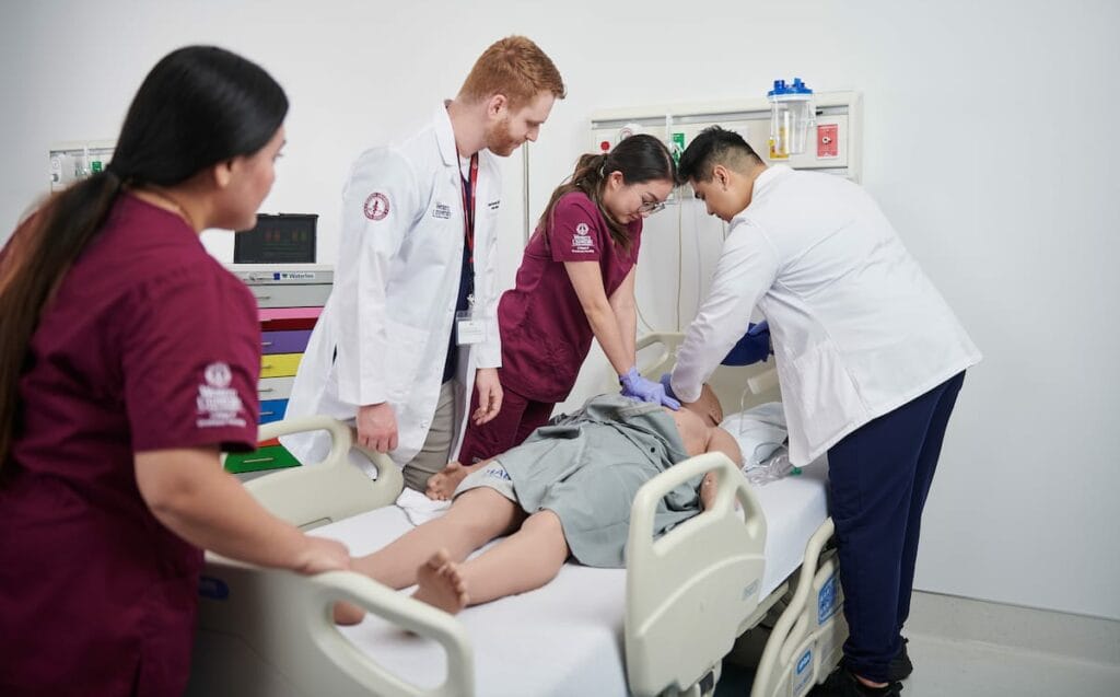 Medical students and instructors practice CPR on a mannequin in a clinical skills lab.