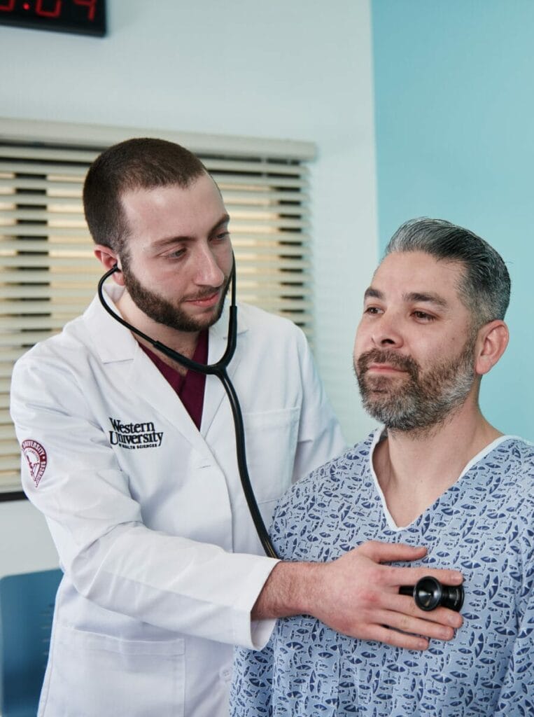 A doctor in a white coat uses a stethoscope to listen to a patient's chest in a medical examination room. The patient is wearing a hospital gown.