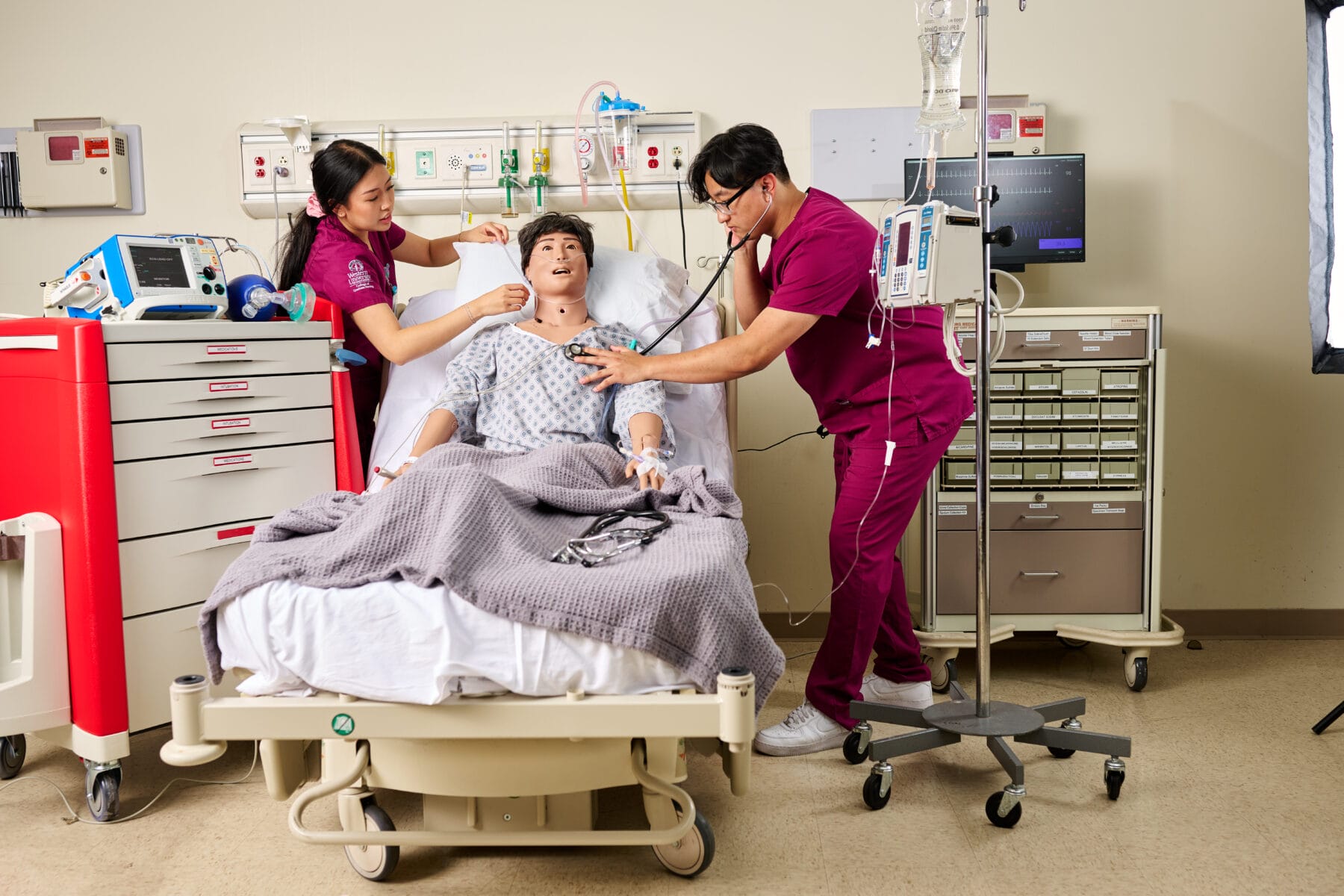 Two medical professionals practice patient care on a hospital bed mannequin, using medical instruments in a clinical training room.