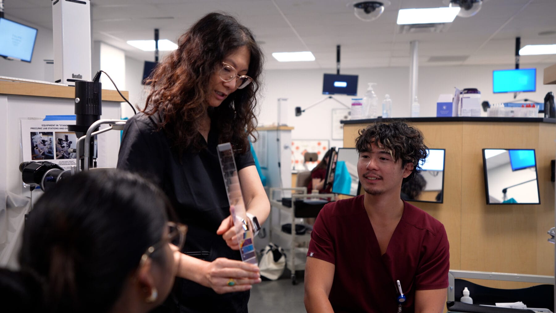 A woman demonstrates a medical tool to two individuals in a clinical setting. They are all wearing scrubs and appear engaged in the discussion.