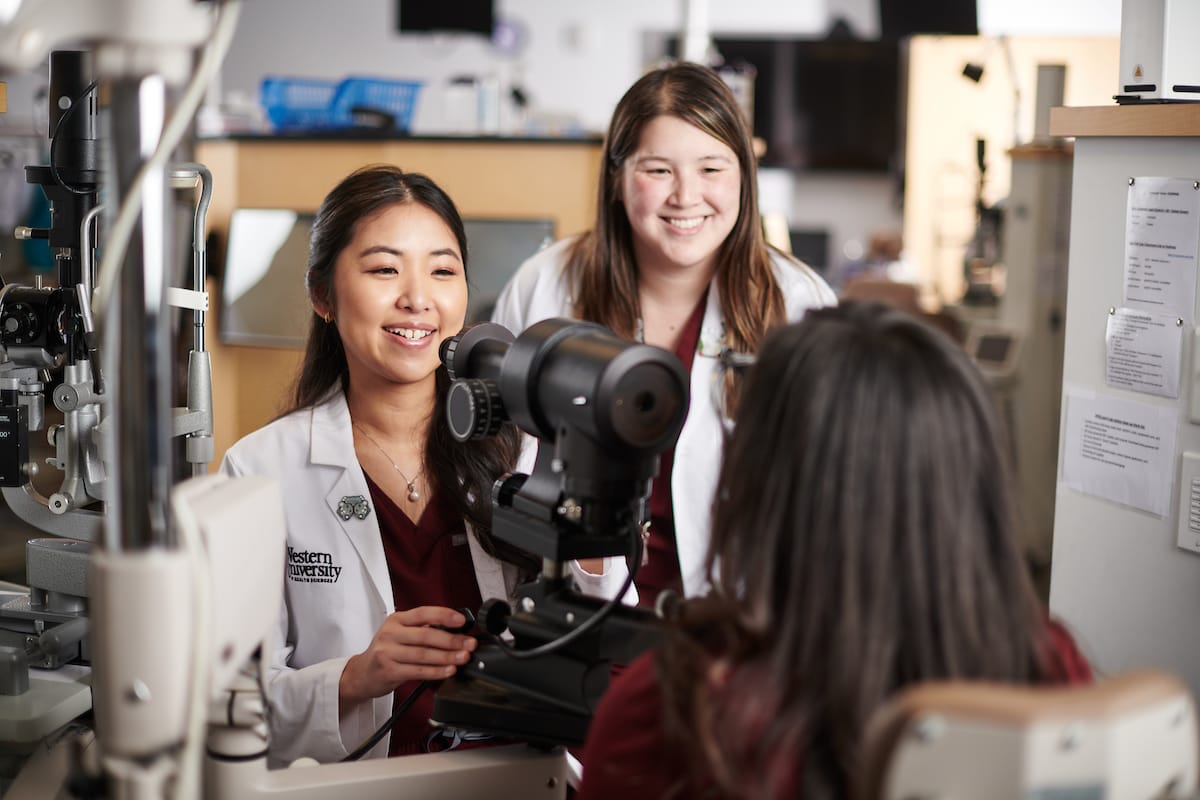 Two female healthcare professionals in lab coats assist a patient during an eye examination with specialized equipment in a clinical setting.