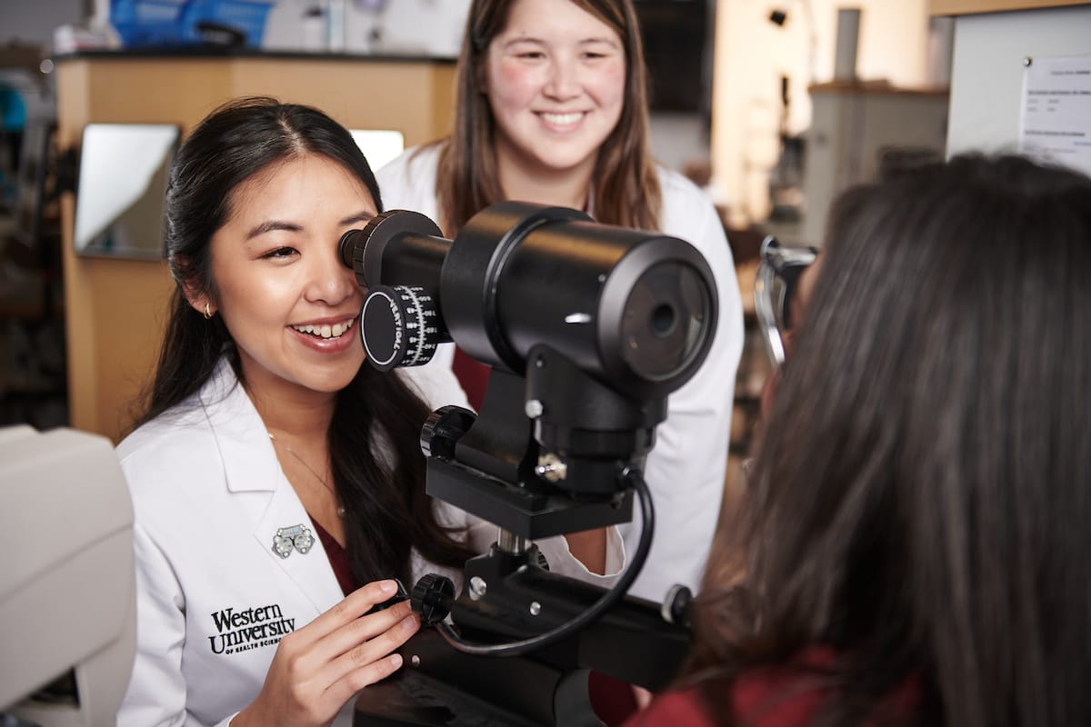 Two women in lab coats, one from Western University, use an optometry device while another woman looks on.