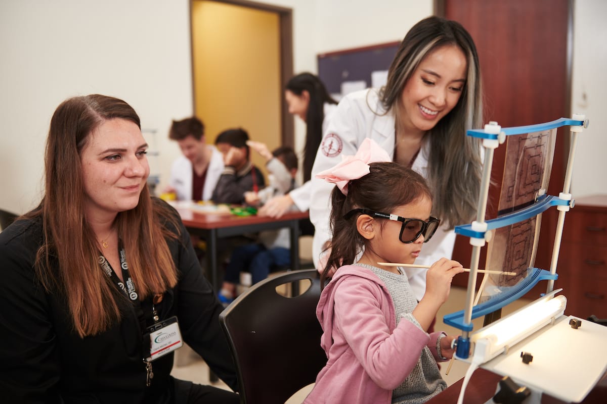 A young girl wearing glasses engages with a sensory toy, assisted by a smiling woman in a lab coat and another woman seated next to her in a room with adults and children.