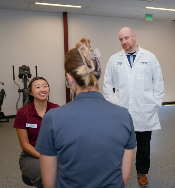 Group of four individuals in a professional setting; one in a lab coat, others in business casual attire. They appear to be in a discussion in a workspace with exercise equipment in the background.