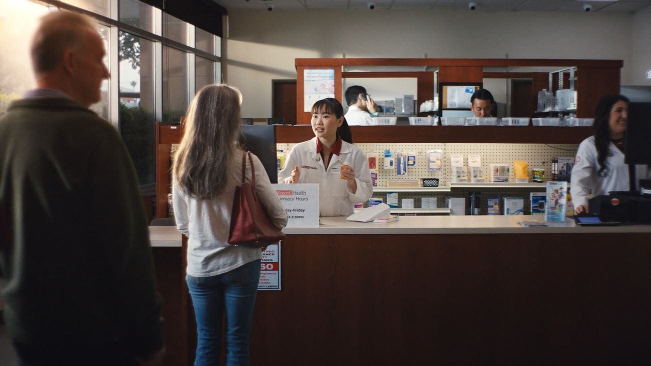 A pharmacist speaks to a customer at a pharmacy counter, with other staff working in the background and brochures displayed on the counter.