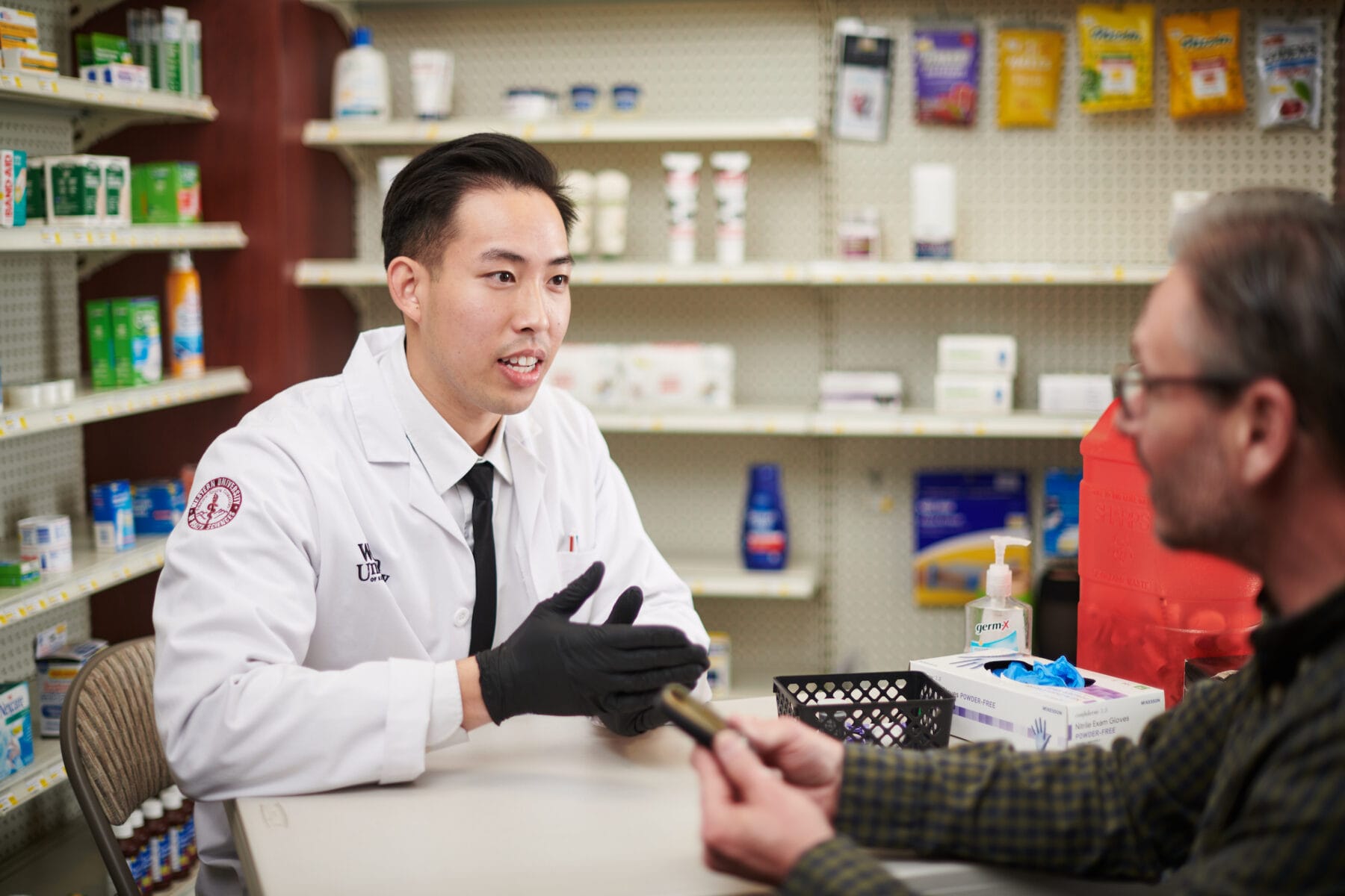 A pharmacist in a white coat and black gloves speaks with a customer seated at a pharmacy counter, surrounded by shelves stocked with various medications and healthcare products.