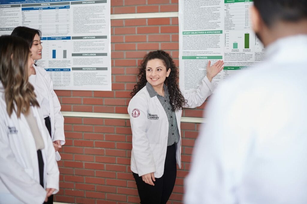 A woman wearing a lab coat presents a scientific poster to a group of colleagues standing beside her.