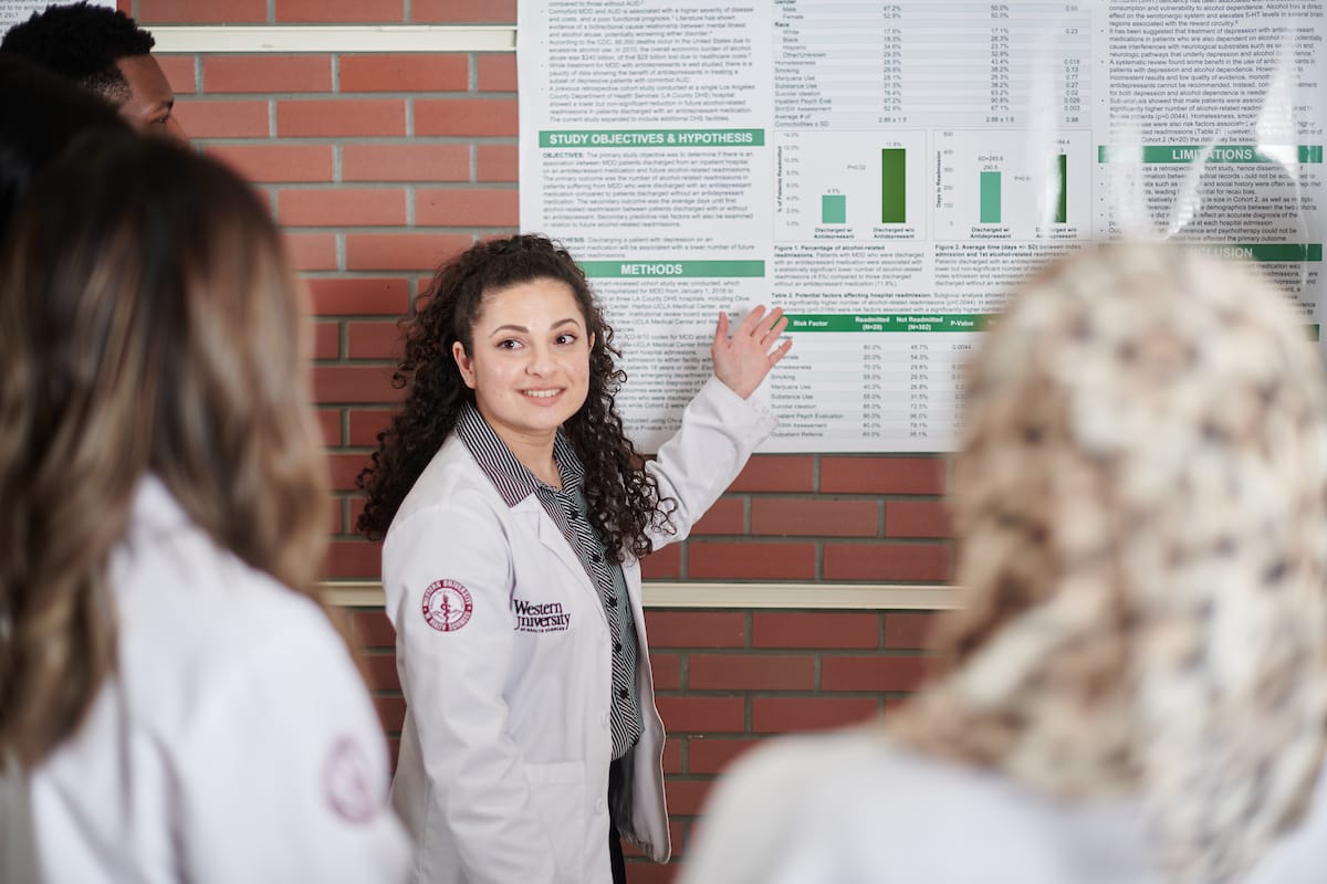 A woman in a lab coat presents research findings on a poster to a group of people, standing in front of a brick wall.