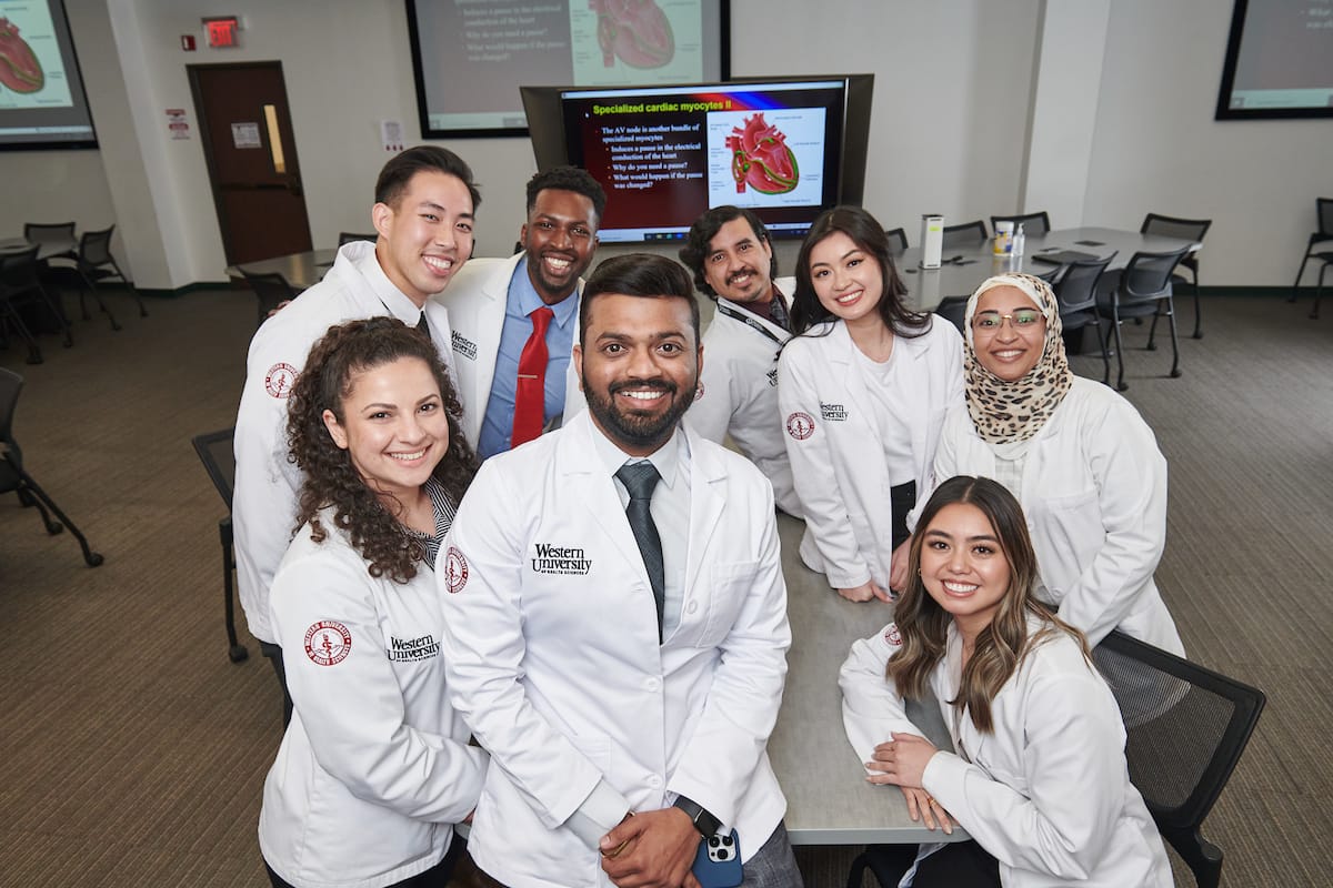 A diverse group of medical students wearing white coats poses for a photo in a classroom with a heart anatomy presentation displayed on the screen behind them.