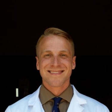 Person smiling in front of a dark background, wearing a white coat and a tie.