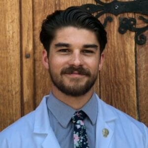 Man with a beard wearing a white coat and floral tie, standing in front of a wooden wall.