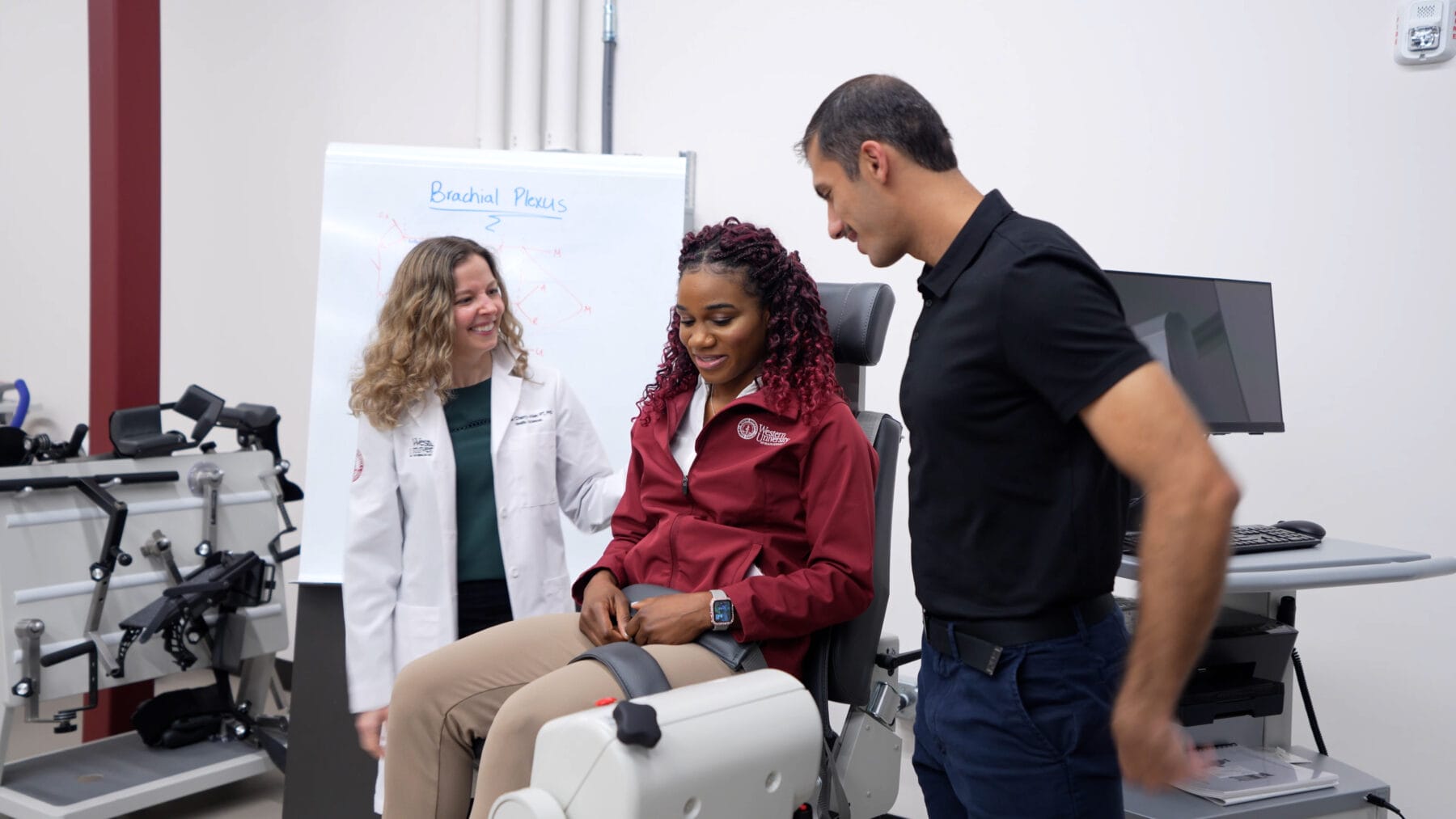 Three people in a medical setting. One woman in a doctor's coat, another woman seated on a medical device, and a man standing next to her. There's a whiteboard and medical equipment in the background.