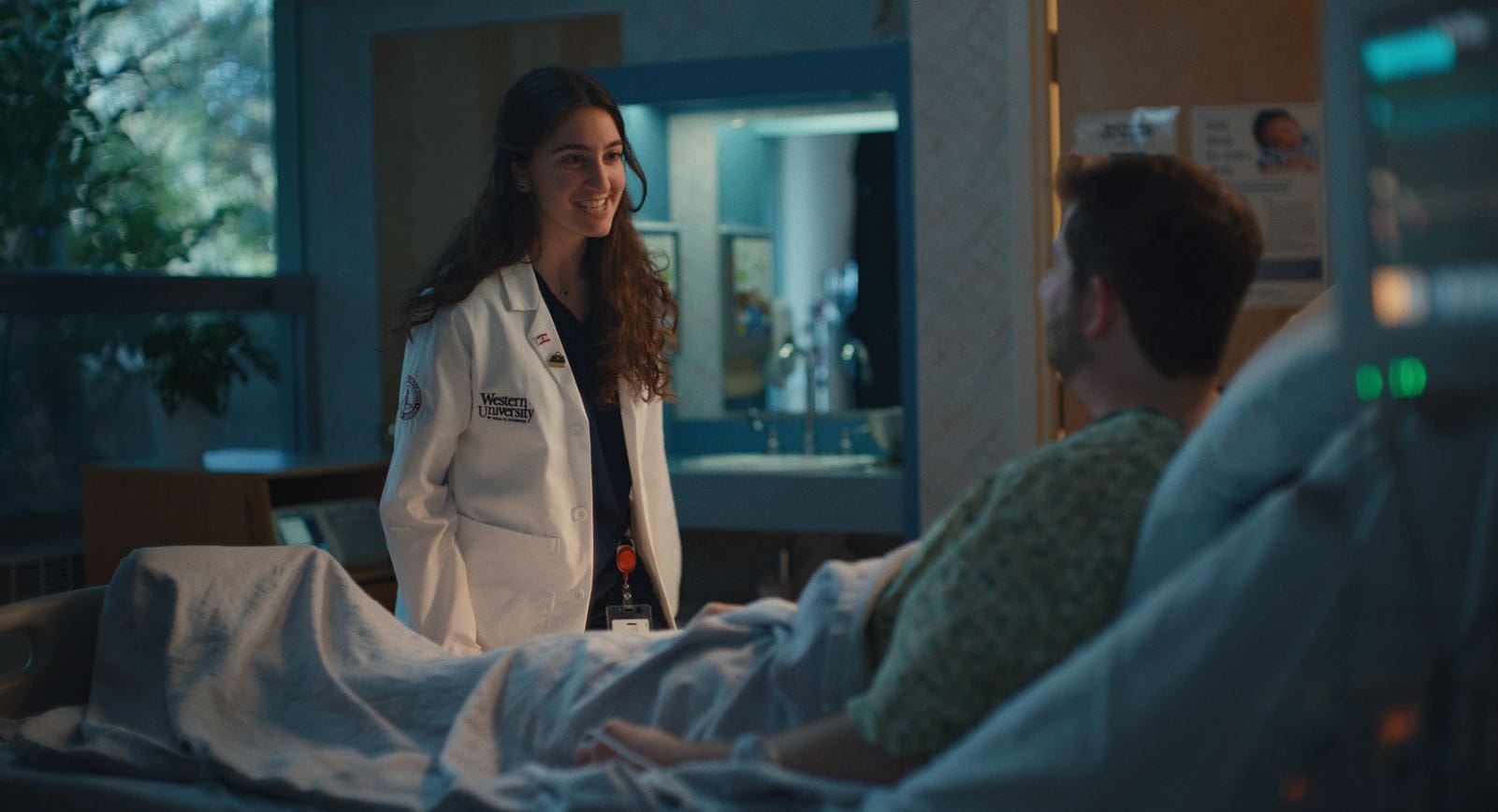 A medical professional in a white coat stands beside a patient lying in a hospital bed, smiling and engaging in conversation.