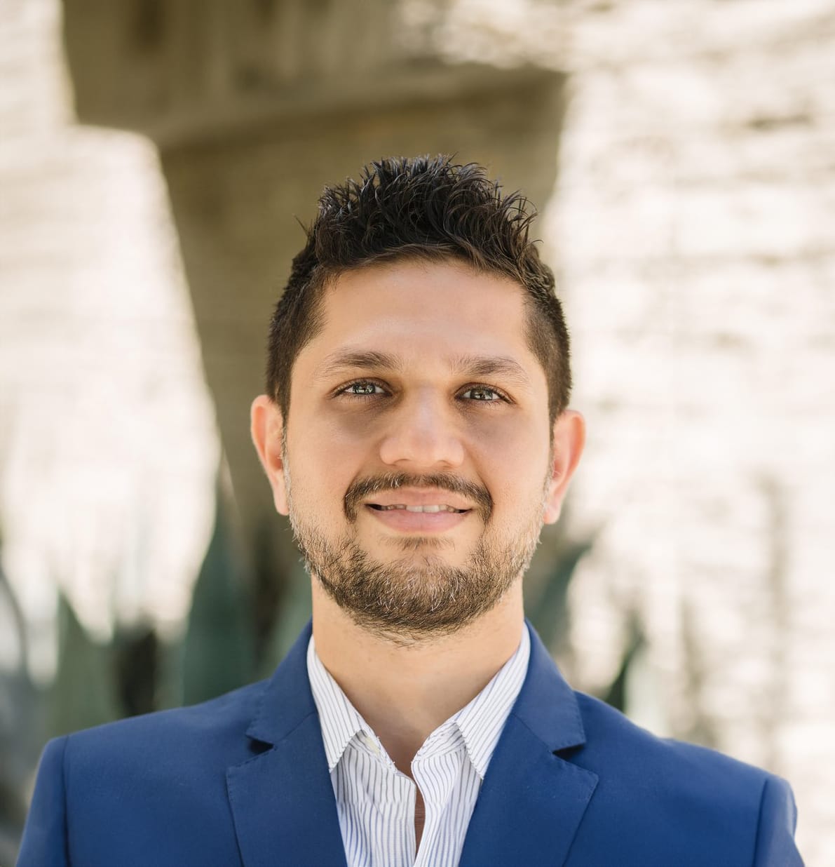 A person with short dark hair and a beard is wearing a blue blazer and striped shirt, standing outdoors with a blurred stone background.